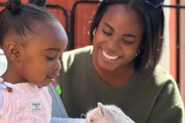 Child in pink overalls sits with a goat while a smiling woman watches.