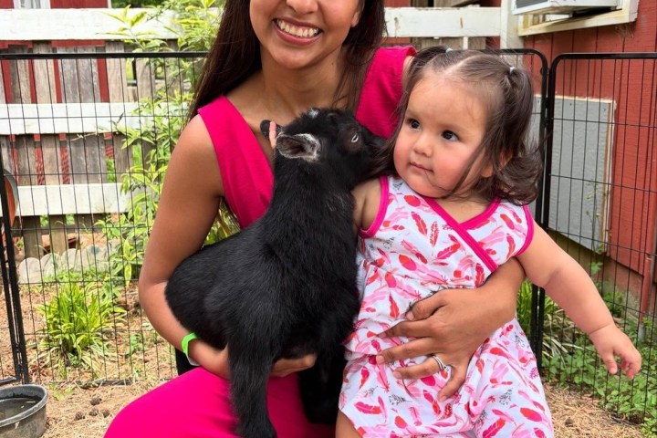 Woman and toddler smiling with a black goat on lap outdoors, near a red house.