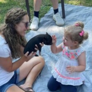 Woman and toddler sitting on a blanket, holding a small piglet on a sunny day.