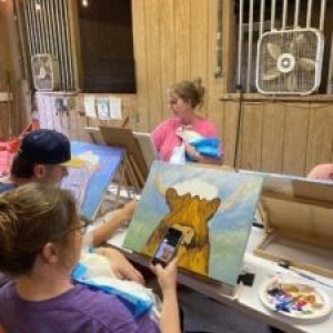 People paint cow portraits in a wooden room with fans.