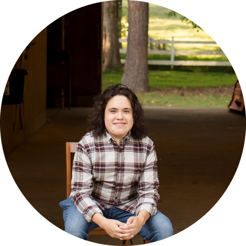 imgi_27_Rosero-05-modified-1024×1024 Person in plaid shirt sitting outdoors in front of a wooden structure and trees.