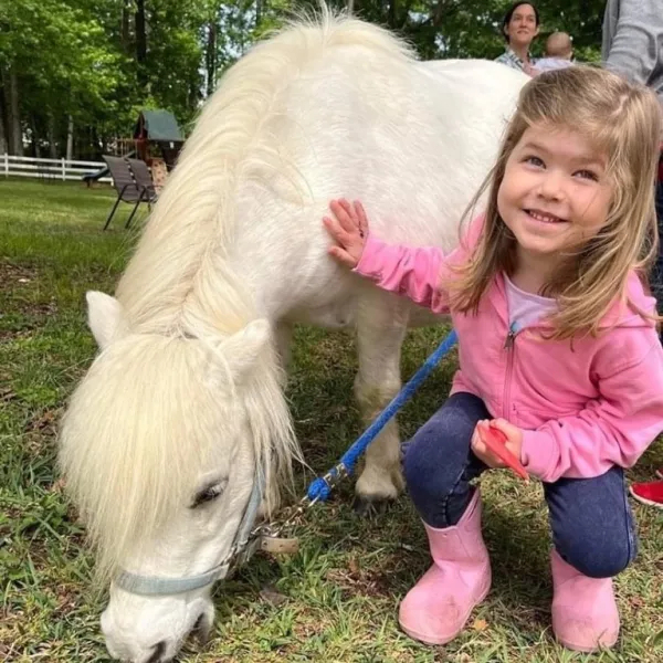 Smiling child in pink jacket and boots petting a small white pony on grass.
