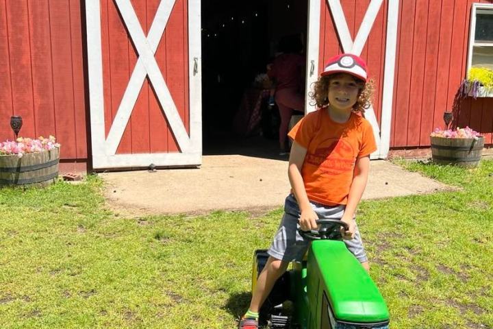Child on a toy green tractor in front of a red barn with white doors.