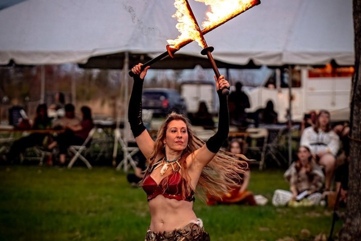 Woman performing fire dance with torches in outdoor setting with large tent and seated audience.