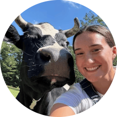 cropped_circle_image (9) Person smiling next to a black and white cow outdoors under a blue sky.
