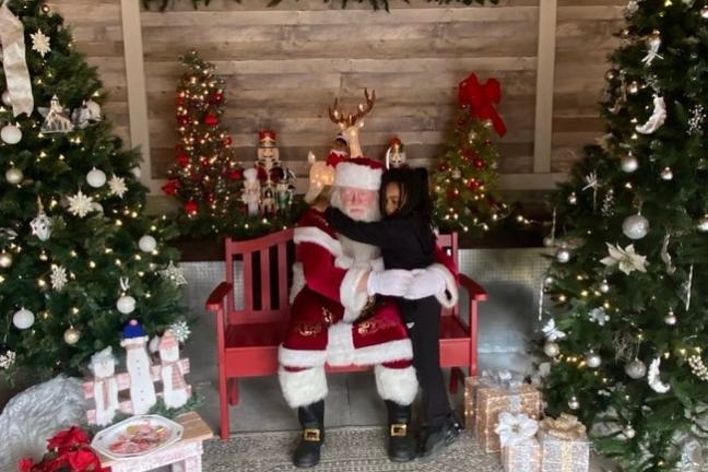 Child hugging Santa Claus on a red bench surrounded by Christmas trees and decorations.