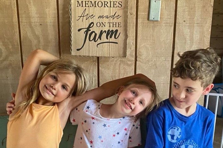 Three kids sitting under a sign that says 'The best memories are made on the farm.'