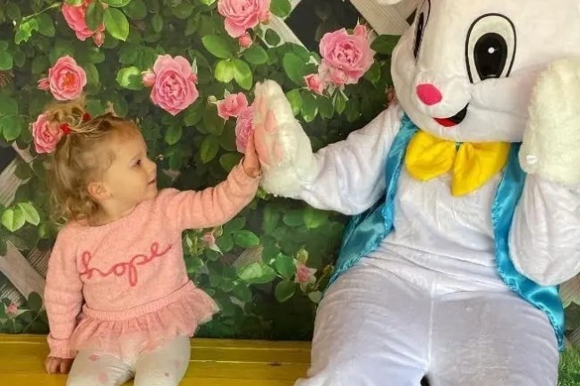 Child in pink outfit high-fives person in Easter bunny costume against floral backdrop.