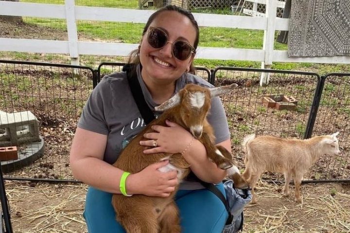 Person seated, smiling, holding a small goat on lap, with another goat nearby.